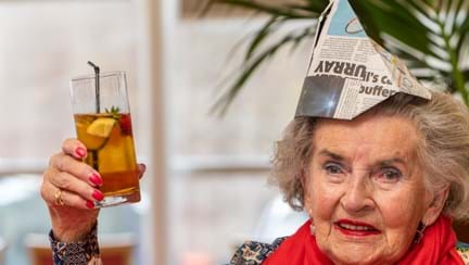 A blind veteran wearing a party hat, holding up a beverage and smiling