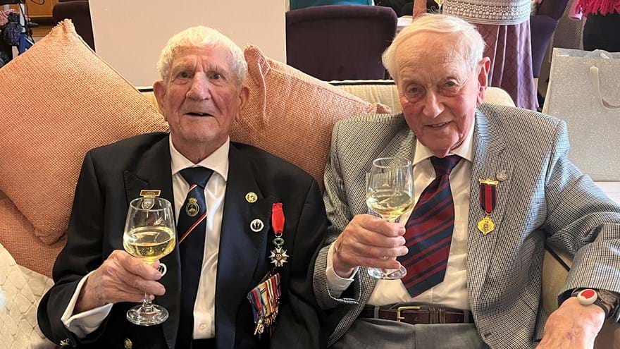 George is sitting next to his friend on a sofa, they are both wearing blazer and medals and toasting the camera with their wine glasses