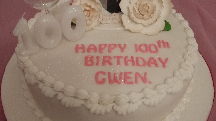 A cake with white frosting, 100th birthday candles, floral detail and a picture of blind veteran Gwen placed on top