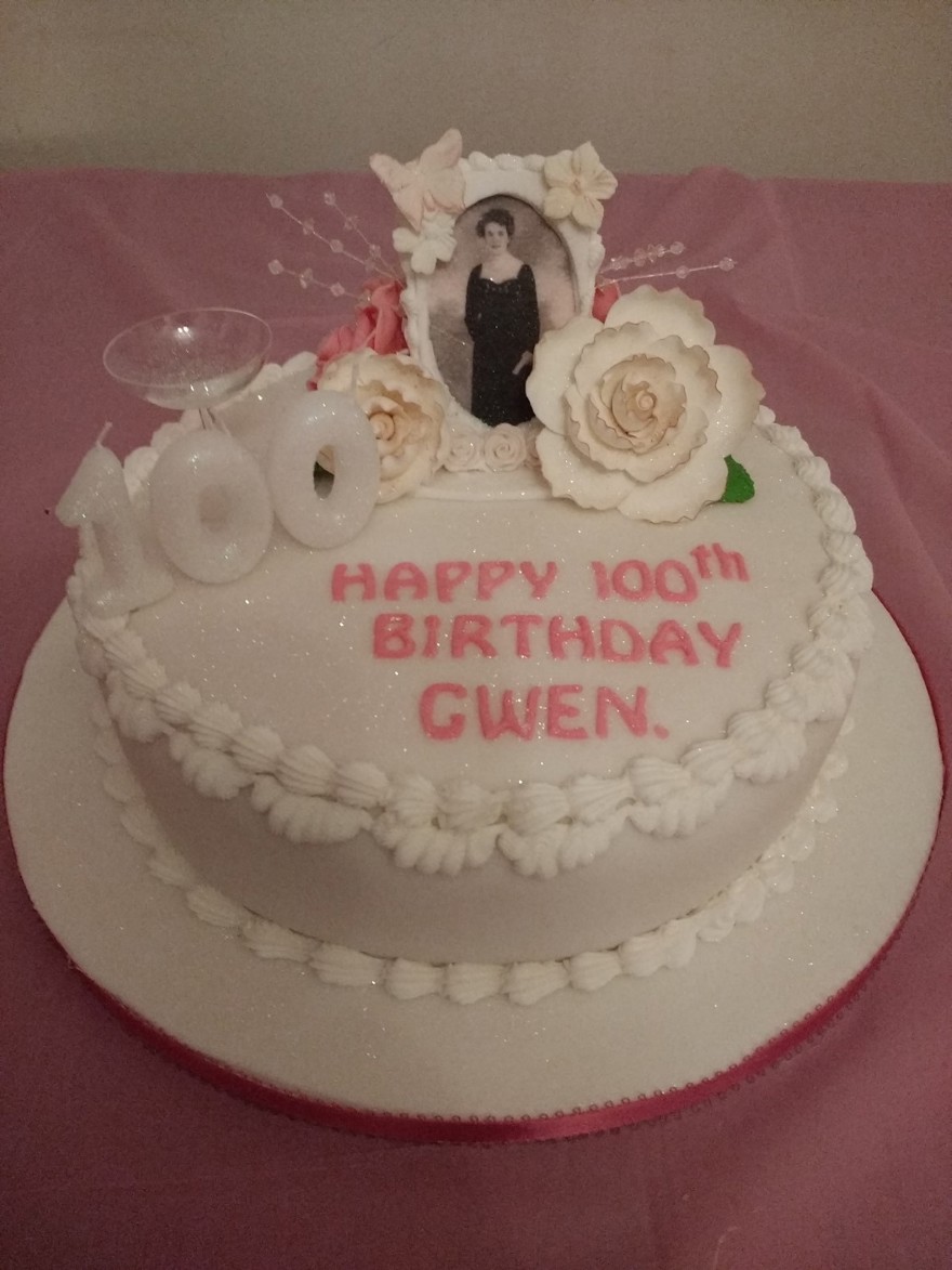 A cake with white frosting, 100th birthday candles, floral detail and a picture of blind veteran Gwen placed on top