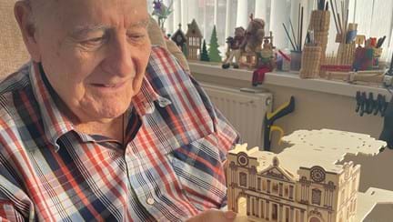 Blind veteran Maurice is looing at his model creation and smiling while sitting in a chair