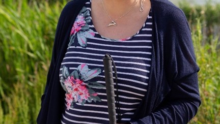 Blind veteran Christine, wearing dark sunglasses and holding a guide cane, as she smiles for a photograph in front of tall grass and a lake in the background