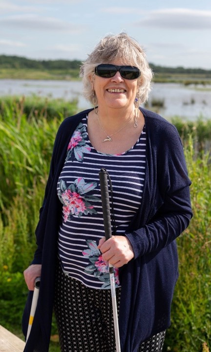 Blind veteran Christine, wearing dark sunglasses and holding a guide cane, as she smiles for a photograph in front of tall grass and a lake in the background