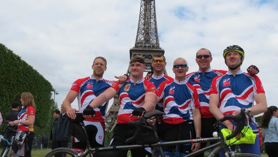 Group of six cyclists in Blind Veterans UK cycling tops posing with a tandem in front of the Eiffel Tower.