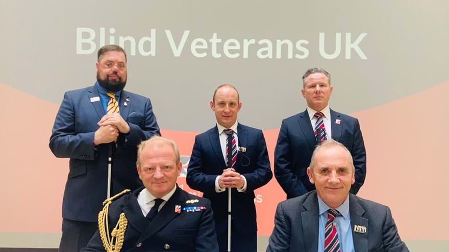 Commodore Bellfield and Nick Caplin sitting at a table signing the Armed Forces Covenant, joined by blind veterans who are standing behind them.