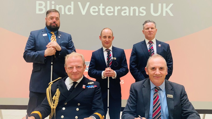 Commodore Bellfield and Nick Caplin sitting at a table signing the Armed Forces Covenant, joined by blind veterans who are standing behind them.