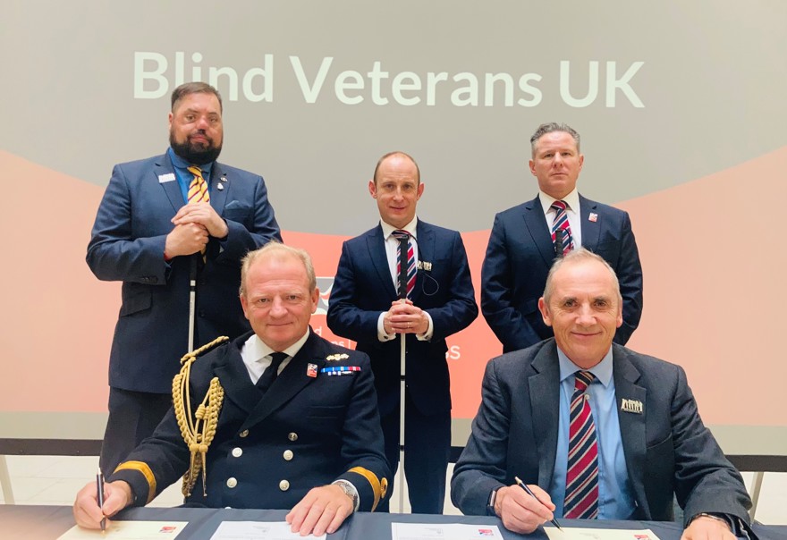 Commodore Bellfield and Nick Caplin sitting at a table signing the Armed Forces Covenant, joined by blind veterans who are standing behind them.