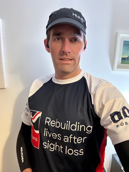 Chris is stood inside his house wearing his Blind Veterans UK running top and a baseball cap