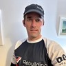Chris is stood inside his house wearing his Blind Veterans UK running top and a baseball cap