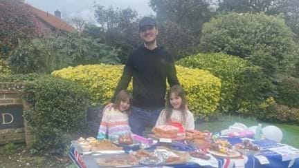 Mollie, Poppy and Will stand behind the table with cakes and Blind Veterans UK flags