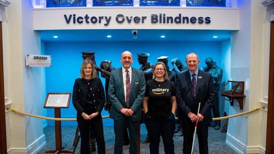 Blind Veterans UK and Amazon staff join a blind veteran standing in front of a sign that reads 'Victory over Blindness'