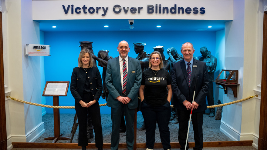 Blind Veterans UK and Amazon staff join a blind veteran standing in front of a sign that reads 'Victory over Blindness'