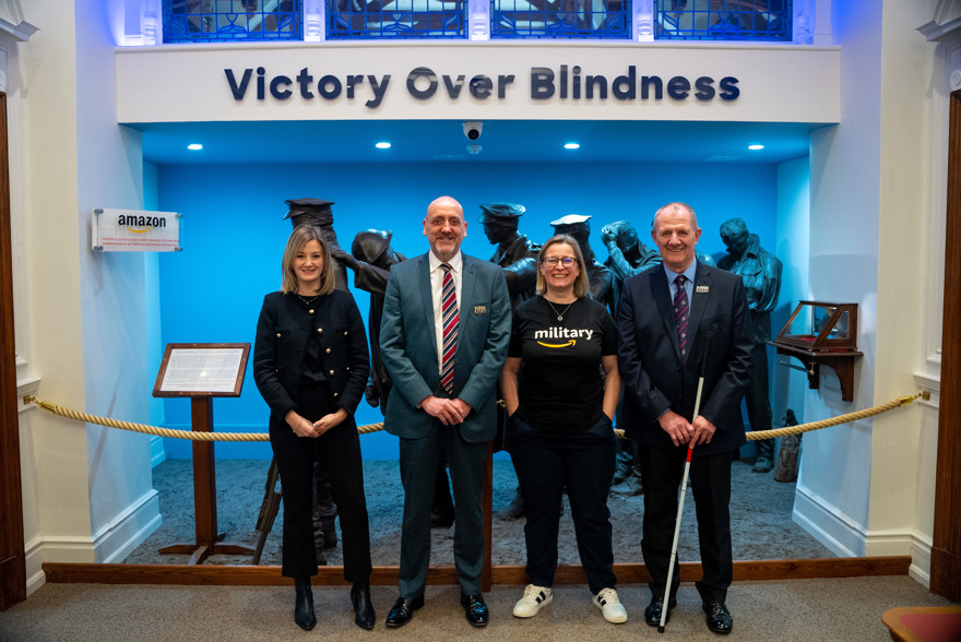 Blind Veterans UK and Amazon staff join a blind veteran standing in front of a sign that reads 'Victory over Blindness'