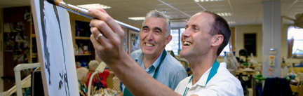 A smiling blind veteran, painting with support from a member of staff