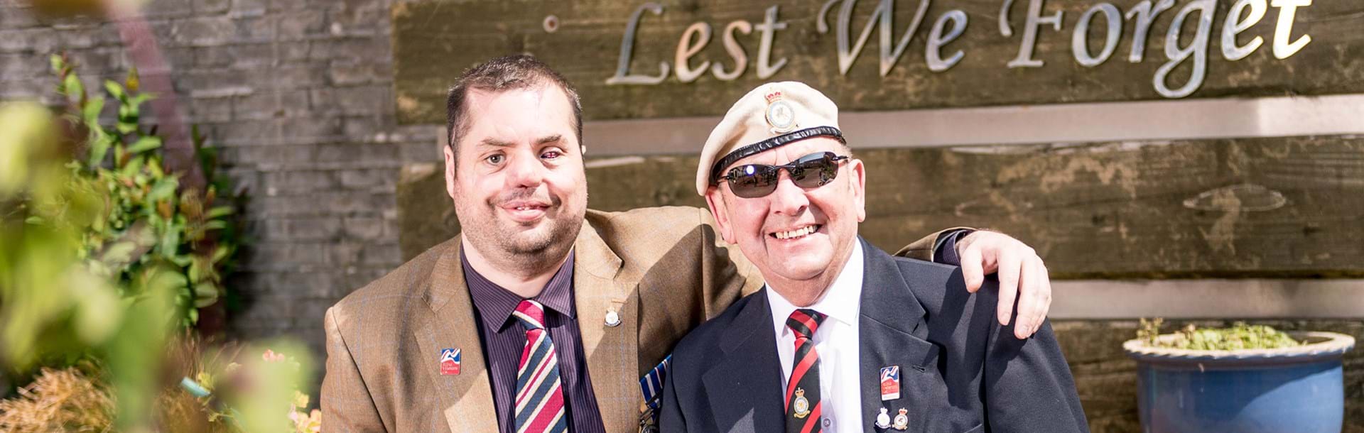 Blind veteran Simon pictured with his arm around fellow blind veteran Tony, they're both smiling as they sit in front of a sign that reads "Lest We Forget"