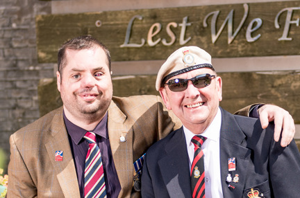 Blind veteran Simon pictured with his arm around fellow blind veteran Tony, they're both smiling as they sit in front of a sign that reads "Lest We Forget"