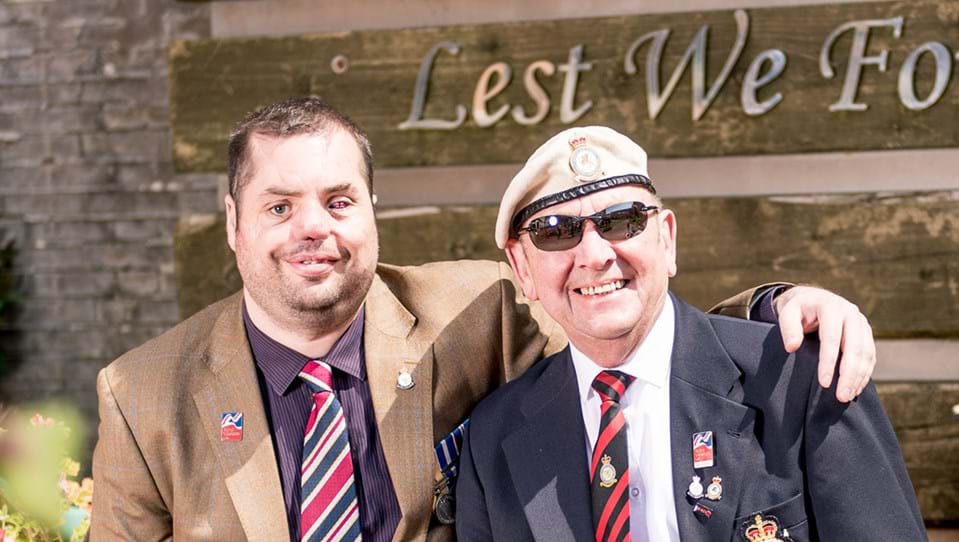 Blind veteran Simon pictured with his arm around fellow blind veteran Tony, they're both smiling as they sit in front of a sign that reads "Lest We Forget"