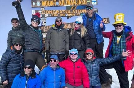 Ian alongside the other climbers all smiling under the Mount Kilimanjaro peak sign