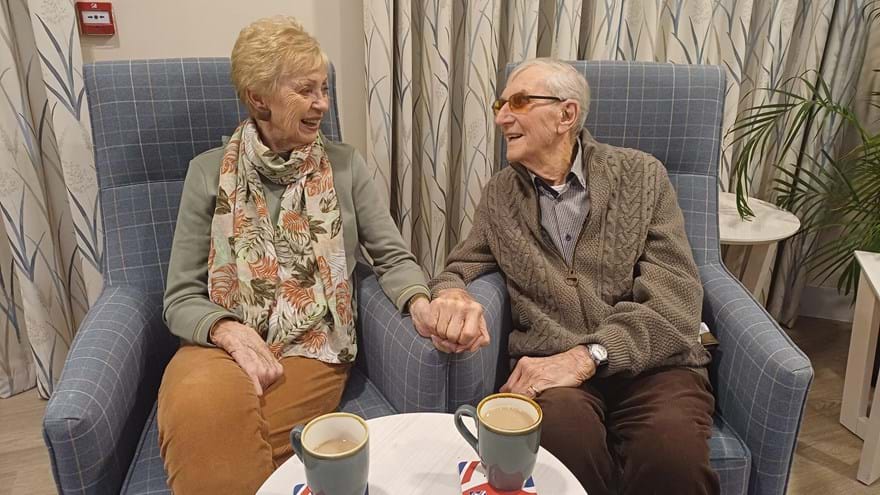 Blind veteran Jim and his wife Elsa sit on arm chairs next to each as they hold hands and look at one another with loving smiles