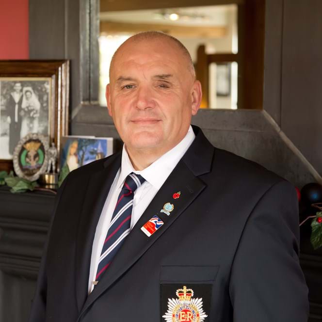 Blind veteran Alan wearing military badges pinned to a smart suit, standing in front of a fireplace decorated with holly.