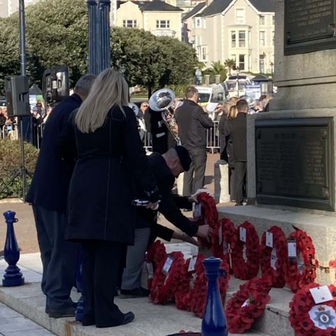 Billy dressed in smart clothes, bending over to lay his poppy wreath beside others. A military band is playing in the background.