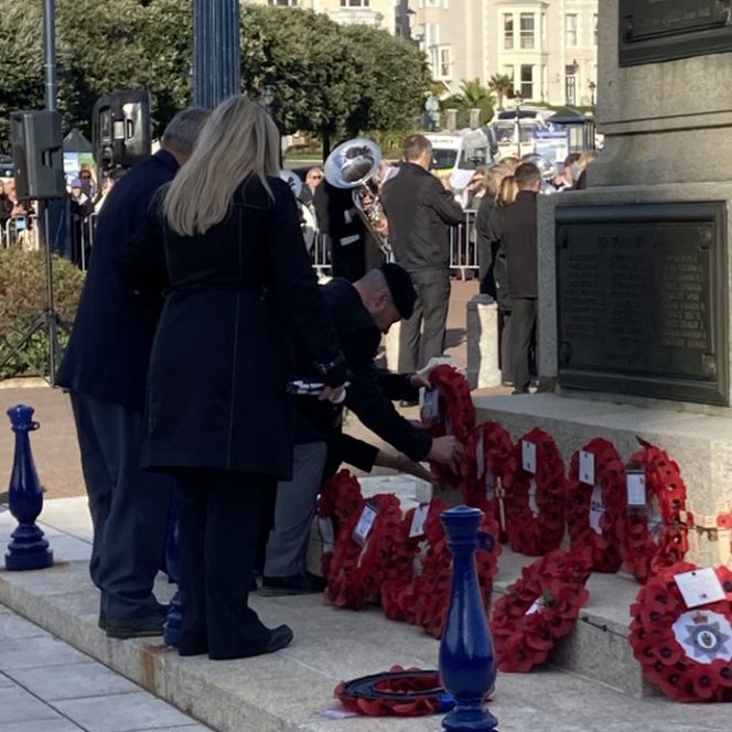 Billy dressed in smart clothes, bending over to lay his poppy wreath beside others. A military band is playing in the background.