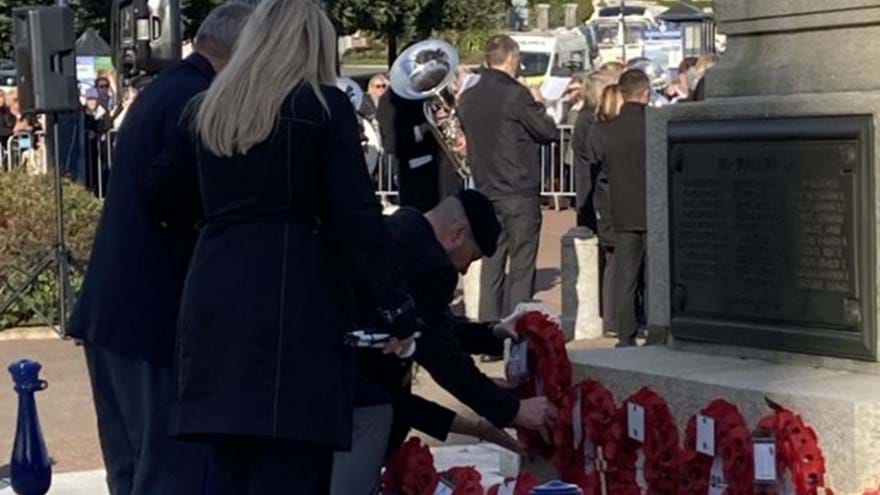 Billy dressed in smart clothes, bending over to lay his poppy wreath beside others. A military band is playing in the background.