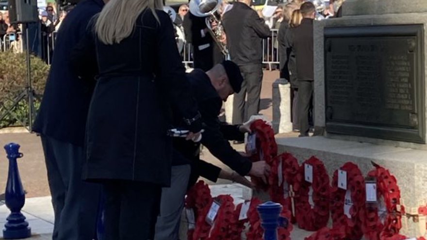 Billy dressed in smart clothes, bending over to lay his poppy wreath beside others. A military band is playing in the background.