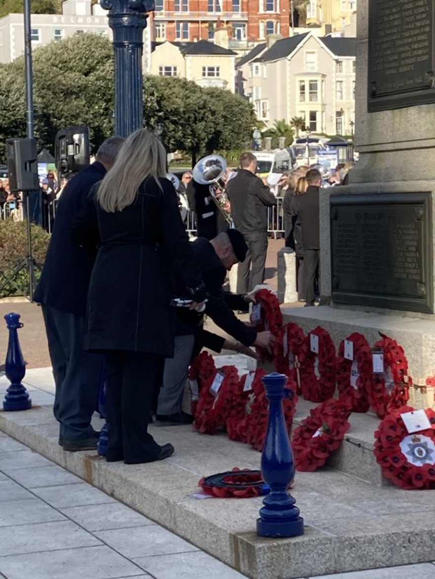 Billy dressed in smart clothes, bending over to lay his poppy wreath beside others. A military band is playing in the background.