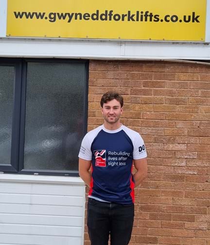 James stood wearing his Blind Veterans UK t shirt in front of a sign for Gwynedd Forklifts