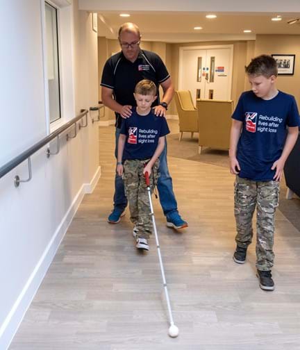 Sammy and Oakley in their Blind Veterans UK t shirts holding a white cane and tightly closing their eyes as they are guided along the corridor at the Rustington Centre