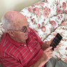 Blind veteran Ron, sitting on a sofa, holding a phone with synapptic software. The screen is displaying a large text main menu.