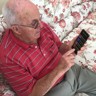 Blind veteran Ron, sitting on a sofa, holding a phone with synapptic software. The screen is displaying a large text main menu.