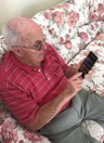 Blind veteran Ron, sitting on a sofa, holding a phone with synapptic software. The screen is displaying a large text main menu.