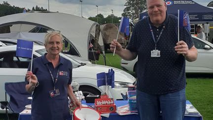 Angela and Tony waving flags in front of a table covered in a Blind Veterans UK table cover