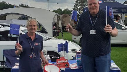 Angela and Tony waving flags in front of a table covered in a Blind Veterans UK table cover 