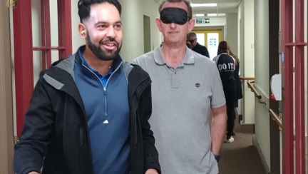 A man wearing a blindfold is holding the arm of another man while he is guided around the inside the the Llandudno Centre