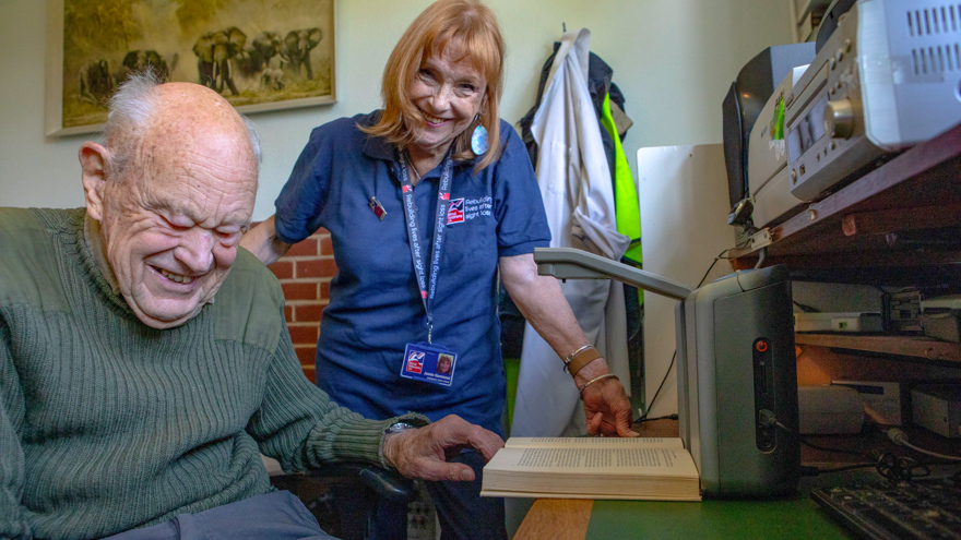 Jennie and Mike are both laughing and looking into camera as Mike puts a book under the reading device