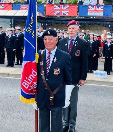 Blind veteran wearing cap and blazer holding the Blind Veterans UK standard