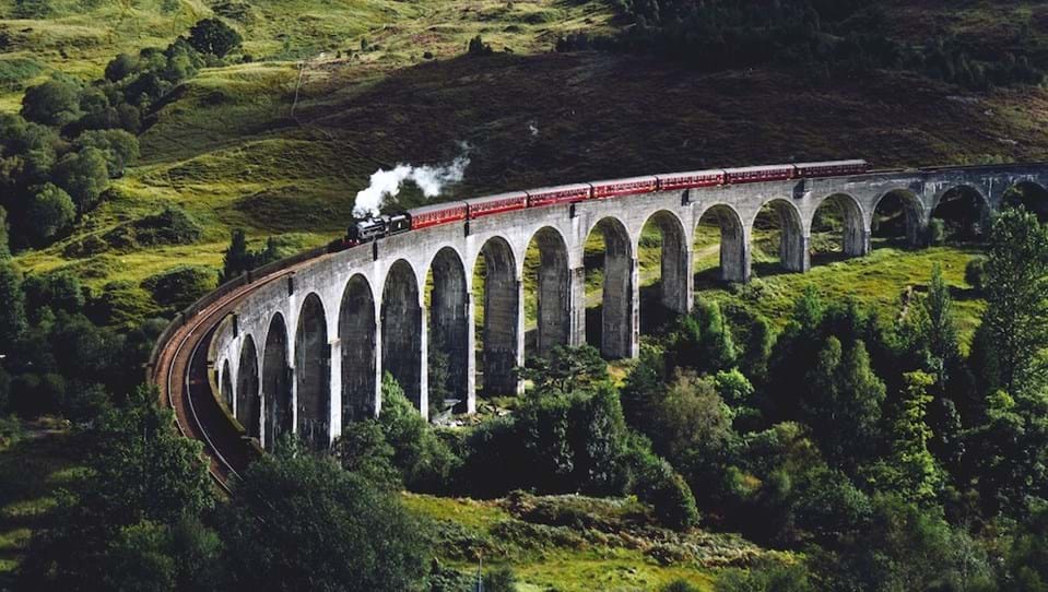 A steam train travelling over the Glenfinnan Viaduct, overlooking green fields and hills.