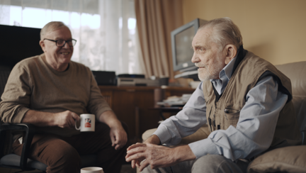 Volunteer Liam is holding a Blind Veterans UK mug as he smiles at blind veteran Ken, both are sitting down in Ken's living room