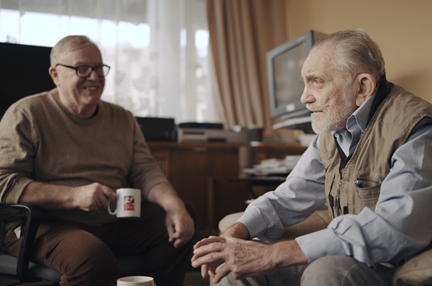 Volunteer Liam is holding a Blind Veterans UK mug as he smiles at blind veteran Ken, both are sitting down in Ken's living room