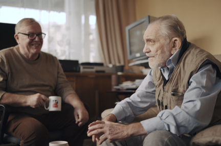 Volunteer Liam is holding a Blind Veterans UK mug as he smiles at blind veteran Ken, both are sitting down in Ken's living room