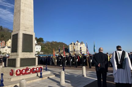 Many people gathered around the Llandudno War Memorial during Remembrance. Poppy wreaths are laid down against the memorial.