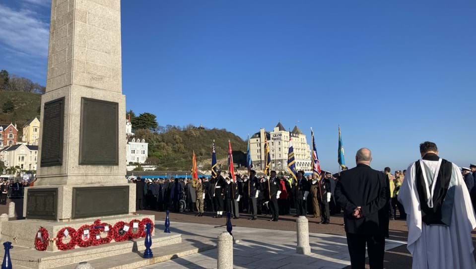 Many people gathered around the Llandudno War Memorial during Remembrance. Poppy wreaths are laid down against the memorial.