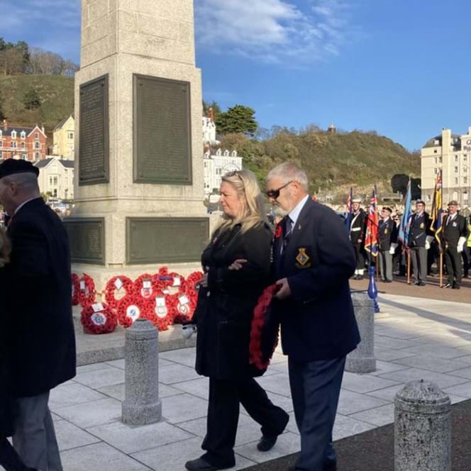 Bob wearing a suit and dark glasses is stood in front of the Llandudno War Memorial waiting to lay a wreath alongside those already placed 
