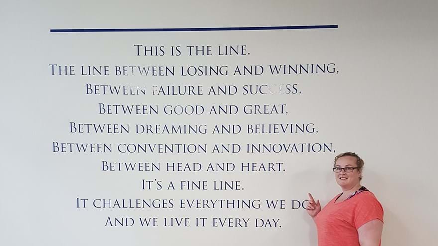 A photo of blind veteran Clare standing next to training equipment and pointing to inspirational message on wall