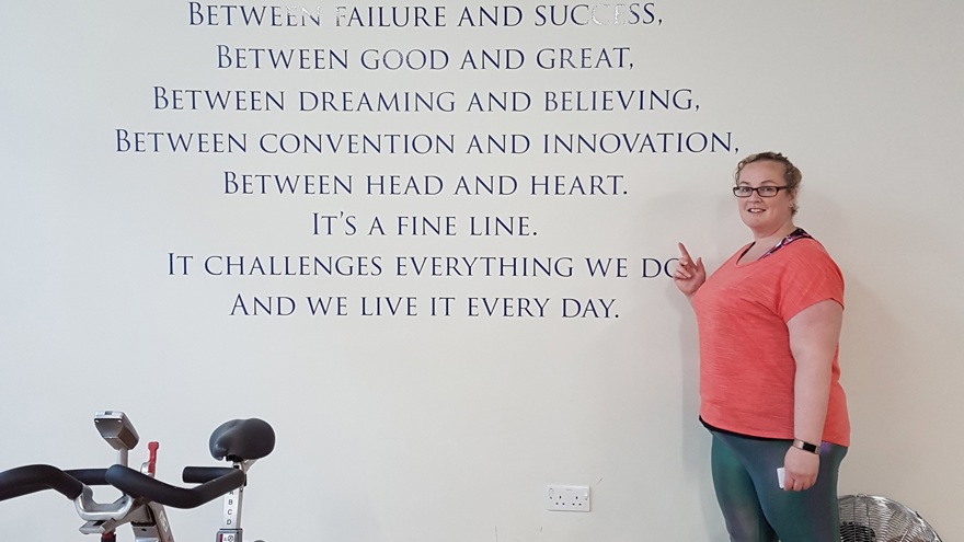 A photo of blind veteran Clare standing next to training equipment and pointing to inspirational message on wall