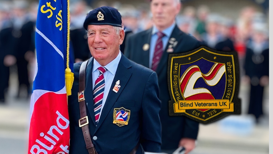 A blind veteran, acting as standard-bearer, dressed in military uniform, holding a Blind Veterans UK flag and wearing a Blind Veterans UK blazer badge
