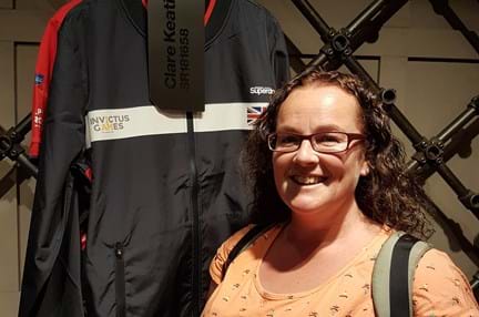 Blind veteran Clare smiling, stood next to her Invictus Games jacket which is hung up behind her